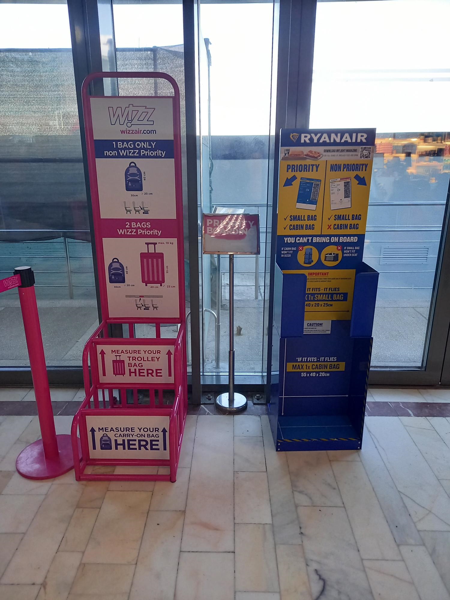Dwa metalowe stojaki do mierzenia bagażu podręcznego w terminalu lotniska: różowy Wizz Air i niebieski Ryanair.
