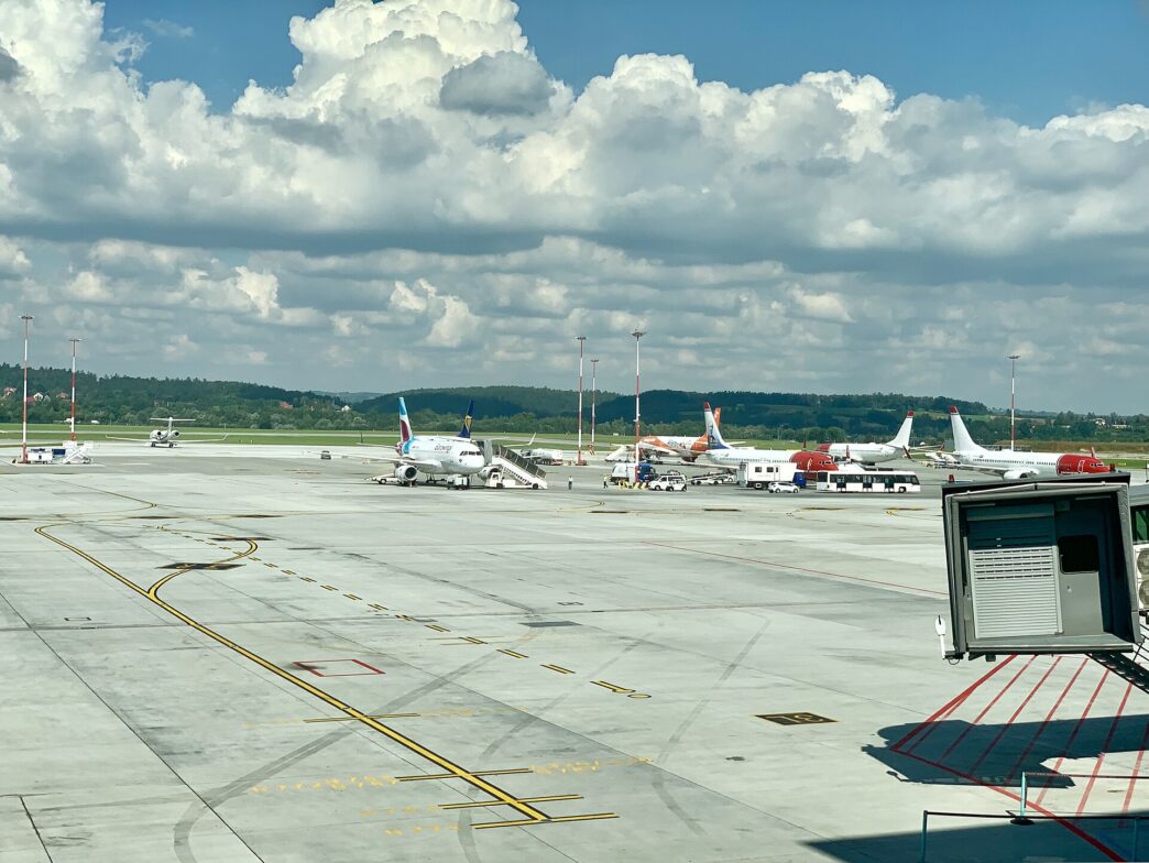 Płyta postojowa lotniska z zaparkowanymi samolotami, rękawem pasażerskim i żółtymi oznaczeniami dróg kołowania