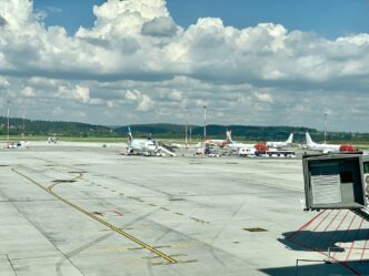 Płyta postojowa lotniska z zaparkowanymi samolotami, rękawem pasażerskim i żółtymi oznaczeniami dróg kołowania