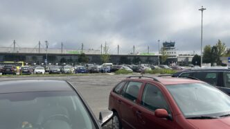 Samochody zaparkowane na lotniskowym parkingu, w tle budynek terminala z napisem Terminal A i wieża kontroli lotów pod zachmurzonym niebem