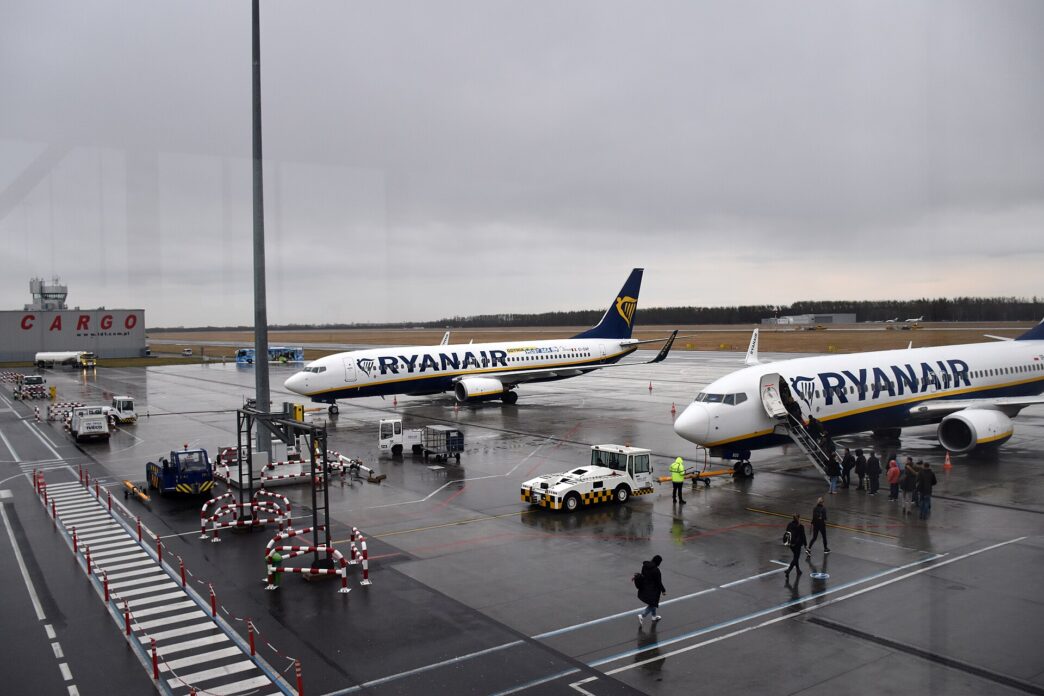 Dwa samoloty Boeing 737 w niebiesko-żółtych barwach Ryanaira na płycie postojowej lotniska, pasażerowie wchodzą po schodach na pokład, obok pojazdy obsługi naziemnej, pochmurne niebo.