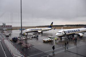 Dwa samoloty Boeing 737 w niebiesko-żółtych barwach Ryanaira na płycie postojowej lotniska, pasażerowie wchodzą po schodach na pokład, obok pojazdy obsługi naziemnej, pochmurne niebo.