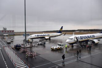 Dwa samoloty Boeing 737 w niebiesko-żółtych barwach Ryanaira na płycie postojowej lotniska, pasażerowie wchodzą po schodach na pokład, obok pojazdy obsługi naziemnej, pochmurne niebo.