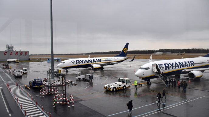 Dwa samoloty Boeing 737 w niebiesko-żółtych barwach Ryanaira na płycie postojowej lotniska, pasażerowie wchodzą po schodach na pokład, obok pojazdy obsługi naziemnej, pochmurne niebo.