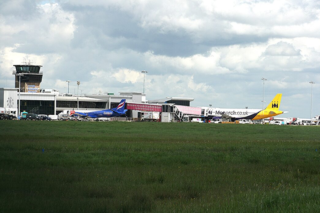 Terminal lotniska Leeds Bradford z wieżą kontroli lotów, przed budynkiem stoją samoloty, na pierwszym planie trawiaste pole
