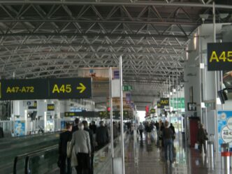 Pasażerowie z bagażem idą korytarzem pirsu A na lotnisku w Brukseli, nad nimi żółte tablice kierunkowe do wyjść A45 i A47–A72