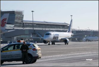 Funkcjonariusz Straży Ochrony Lotniska przy służbowym samochodzie na płycie lotniska, w tle kołujący samolot pasażerski i terminal