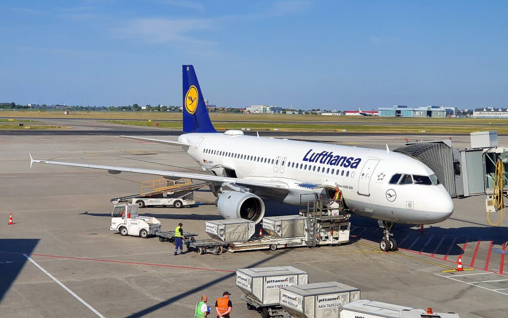 Samolot Airbus A320 w barwach Lufthansy przy rękawie pasażerskim na płycie postojowej, obok wózki z kontenerami bagażowymi i pracownicy obsługi naziemnej