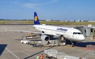 Samolot Airbus A320 w barwach Lufthansy przy rękawie pasażerskim na płycie postojowej, obok wózki z kontenerami bagażowymi i pracownicy obsługi naziemnej