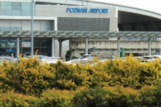 Fasada terminalu pasażerskiego z niebieskim napisem Poznań Airport, pod nim zadaszony podjazd, parking z samochodami i żywopłot na pierwszym planie