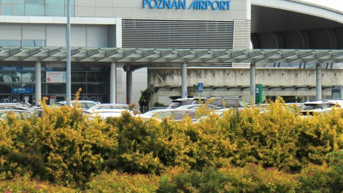 Fasada terminalu pasażerskiego z niebieskim napisem Poznań Airport, pod nim zadaszony podjazd, parking z samochodami i żywopłot na pierwszym planie