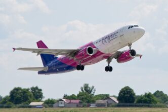 Samolot Airbus A320 w biało-fioletowo-różowych barwach Wizz Air wznosi się nad polami i zabudowaniami