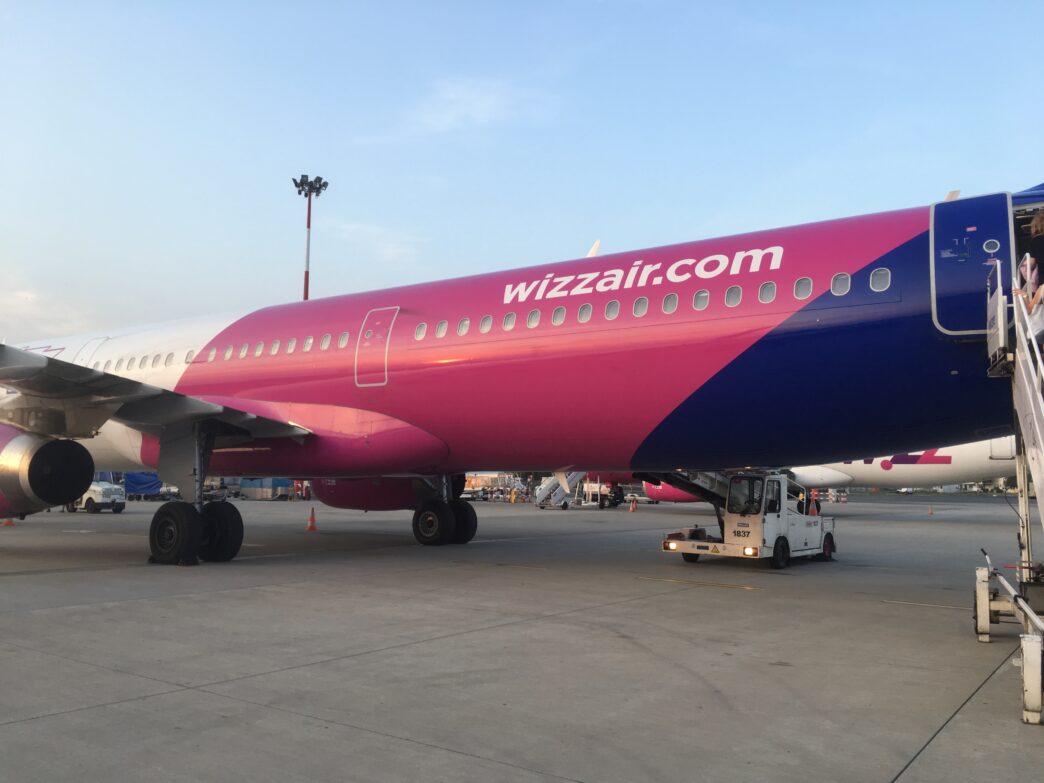 Samolot Wizz Air w różowo-fioletowych barwach stoi na płycie postojowej, obok schody pasażerskie i ciągnik lotniskowy.